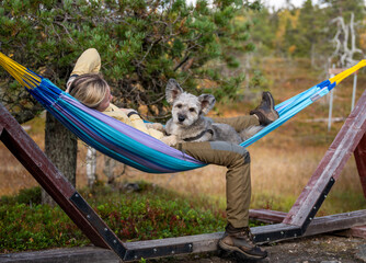 Woman relaxing in hammock with dog wearing outdoor clothing in forest nature