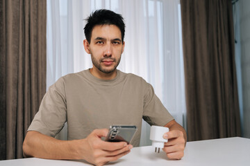 Portrait of young man holding smart socket and smartphone, looking at camera, connecting smart home device for wireless automation, setup and iot control in modern apartment.