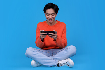 Young Asian male sitting cross-legged and playing mobile game on blue background