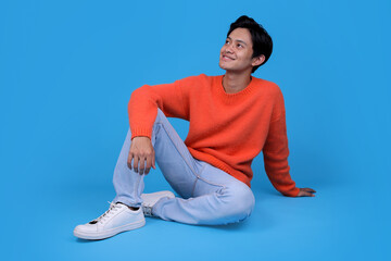 Young man in orange sweater looking to the side and sitting comfortably on blue background