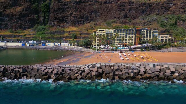 Smooth aerial pan across Calheta Beach and waterfront in Madeira, Portugal. Drone view shows sandy shoreline, breakwater, beachfront hotels, sunbeds, and calm Atlantic waters
