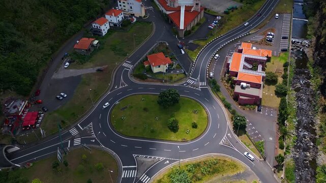 Aerial view of a modern roundabout near S&atilde;o Vicente Valley in Madeira, Portugal, showing cars moving through winding mountain roads, residential buildings, and lush green surroundings