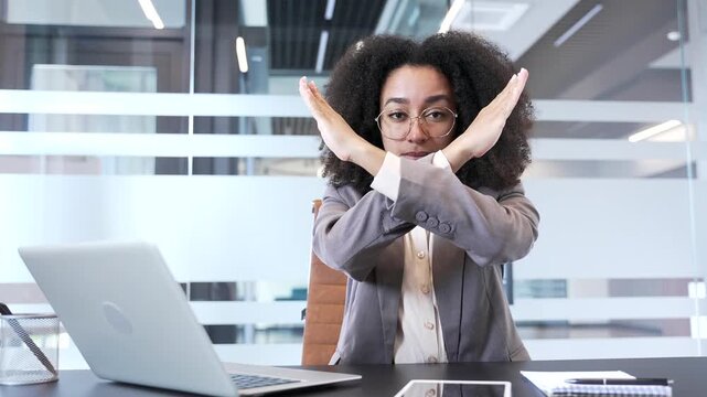 African American businesswoman shaking a no sign with her index finger, showing crossed arms, X sign while sitting in business office. Woman demonstrates a gesture of prohibition, denial, disagreement