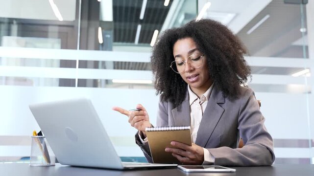 African american businesswoman watching video call online conference taking notes looking at laptop screen in office. Female worker listening remote business training, talking communicates with tutor