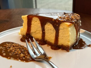 Classic cheesecake slice topped with salted caramel sauce and crunchy nut pieces, served on a white plate in a cozy caf&eacute; setting.