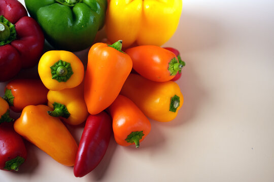a vibrant assortment of red, green, yellow, and orange bell peppers and mini sweet peppers arranged on a smooth, off-white surface highlighting their glossy texture, freshness, edible, capsicum