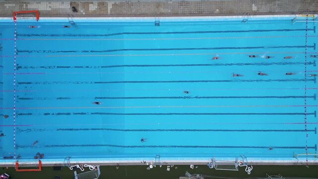 Top-down aerial view of outdoor swimming pool with swimmers moving along lanes. Clean geometric composition with blue water, ideal for sport, fitness, training and summer recreation themes