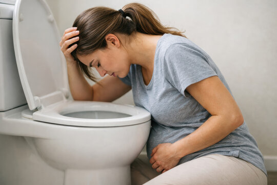 Pregnant woman sitting on bathroom floor feeling nauseous and unwell while vomiting into toilet during early morning sickness