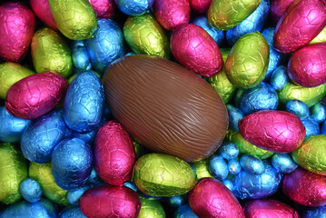 Pile or group of multi colored and different sizes of colourful foil wrapped chocolate easter eggs in blue, pink, red, yellow and lime green with a large brown milk chocolate egg in the middle.