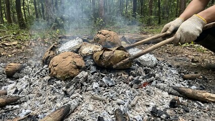 Obraz premium Person wearing gloves using tongs to carefully turn food packets wrapped in foil and leaves, baking in hot ashes and embers from a campfire in a forest
