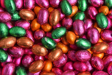 Large & small pink, green, orange and bronze foil wrapped chocolate easter eggs, against a peach orange background.