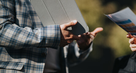 Business interaction outdoors: hands pass documents and a tablet, conveying collaboration and exchange.