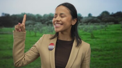 Woman in park smiles with finger raised wearing an i voted badge celebrates outdoors against green park backdrop suggesting civic pride and engagement in the united states.