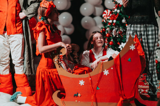 Performers in red costumes gather around a decorative sleigh on a Christmas theater set. Children and adults prepare props and decorations for a holiday stage show.