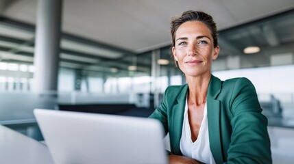 Mature professional secretary or project manager woman in her 40s working on laptop at modern office desk, looking at camera with calm focused expression. Middle aged female corporate employee