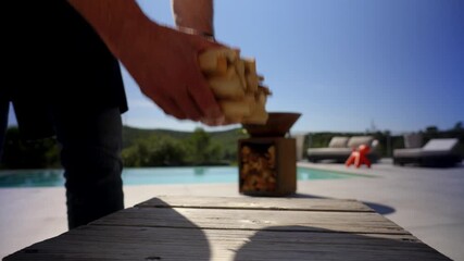 Hands stacking firewood on a table near a poolside fire pit, capturing outdoor prep and relaxed lifestyle