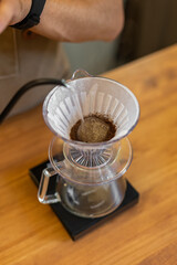 A person pours hot water over coffee grounds in a pour-over coffee maker. The setup includes a glass carafe and a wooden table surface.