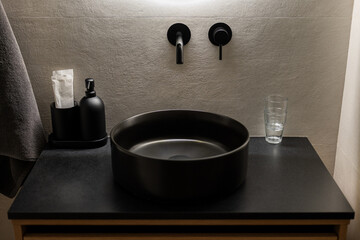 A modern bathroom sink with a round black basin on a dark countertop. There are black wall-mounted faucets, a glass, and a soap dispenser nearby.