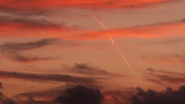An airplane flies through a colorful sunset sky. Contrails are visible against the vibrant clouds, creating a striking contrast with the warm hues.