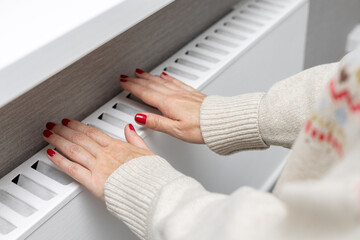 A woman warms her hands by holding them over a radiator. A girl warms herself near a heating radiator. person warming up. thermal energy. heating season. cold season. utility bills. heating tariffs. 