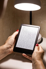  A woman reading an e-book at home, vertical photo. reading an e-book by lamplight. reading a book at the table. woman holding e-book. hand holding ereader. e-ink book. vertical photo