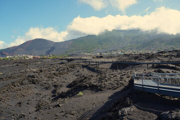 Elevated walkways cross the lava fields of La Palma near the Tajogaite volcano, providing access through the dark volcanic terrain with forested slopes and scattered settlements in the background
