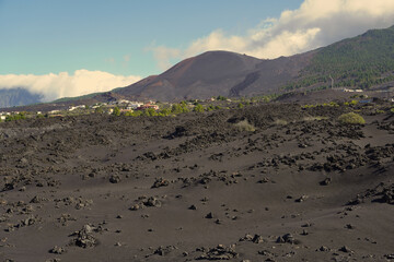 Volcanic terrain on La Palma with black lava fields leading toward the cone of the 2021 Tajogaite volcano. The scene shows rugged formations, nearby settlements and forested slopes under a bright sky