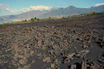 Volcanic landscape on La Palma, showing lava fields and rugged terrain with the green ridges of the island rising in the background under a clear sky. A striking view of recent geological activity