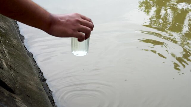 Close up hand collecting water sample from murky river canal into a clear glass to check quality. Concept of water pollution, environmental contamination, lack of clean water, and sanitation crisis.