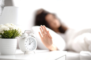 On the bedside table, there is a white retro alarm clock and a potted plant with white flowers; in the background, a person in bed is blurred, reaching out their hand to turn off the alarm. 