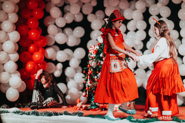 Two girls in red dresses perform a festive dance on a decorated stage. A Christmas theater scene with balloons, a Christmas tree, and holiday costumes creates a joyful ambiance.