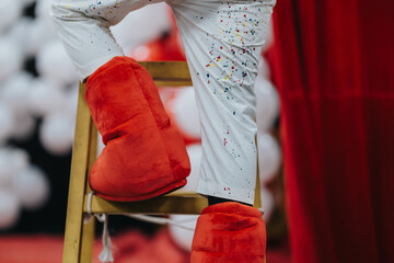 Bright red plush stage boots rest on a wooden ladder. A performer in white paint-splattered pants stands near a red curtain, creating a lively backstage feel.