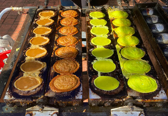 Street snack cooking in circular molds on griddle, showing batter stages and vibrant green pandan variation