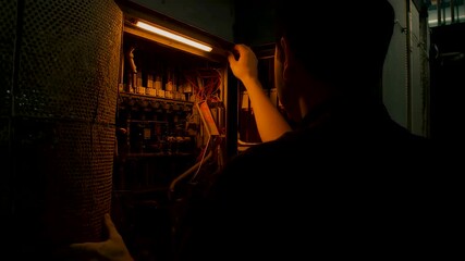 Electrician inspecting control panel wiring inside dimly lit industrial room showing technical maintenance work safety checks and focused expertise