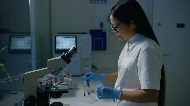 Female lab technician observing a blood sample under the microscope during detailed medical analysis, side view. Blood smear and hematology concepts.