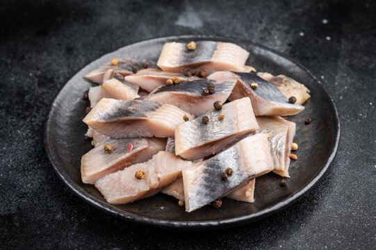 Pickled herring chunks cured in salt and brine served with spices and herbs. Traditional seafood on dark textured background.