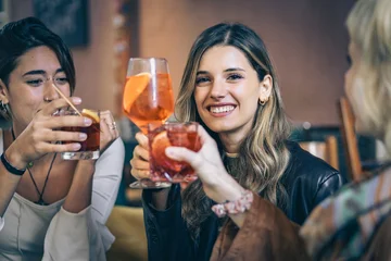 Fototapeten Bar Three young women toasting with cocktails in a bar, friendship and social lifestyle moment  © Lomb