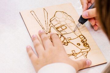 Child carefully uses pyrography tool to burn detailed design on a plywood board indoors. Creative process, focused hands crafting patterns, promoting artistic development and hobby skills for kids.