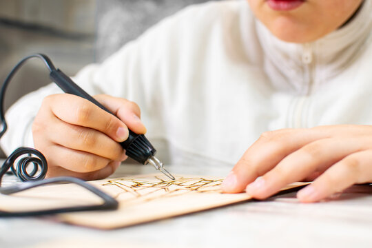 Focused child applies a hot pyrography tool to etch an elaborate illustration on a wooden board at class. Artistic activity, steadily create detailed lines, patient hobby development for children