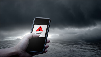 Hand holds a smartphone displaying a tsunami alert message against a dramatic backdrop of dark, ominous clouds and turbulent ocean waters. Technology in disaster warning and emergency preparedness