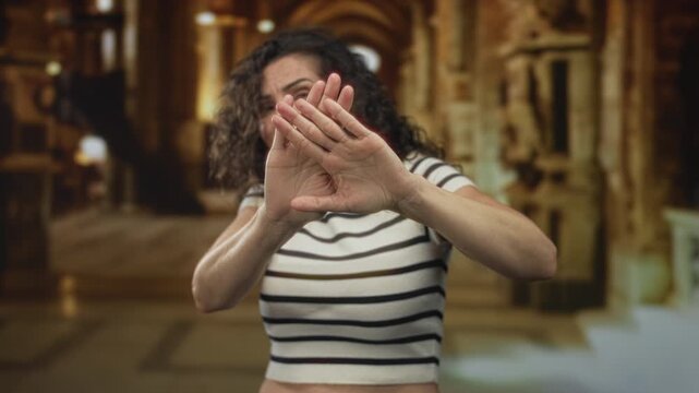 Young hispanic woman blocks view with extended palms covering view partially in golden ornate building; rejection.