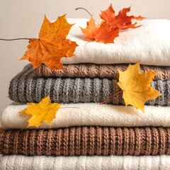 Stacked, folded sweaters in various neutral colors, adorned with vibrant orange and yellow fall leaves on top. A cozy autumn arrangement