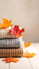 Stacked sweaters and autumn leaves on a light wooden surface against a neutral backdrop, evoking coziness