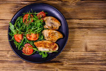 Roasted chicken breasts and salad with arugula and cherry tomatoes in a black plate. Top view