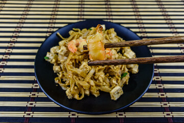 Black plate with pad thai on a bamboo mat. Chopsticks with shrimp