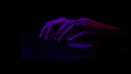 Silhouette of hands typing on a laptop keyboard under dramatic purple and red lighting in darkness