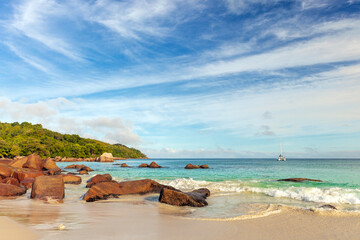 A tranquil tropical beach scene features large reddish rocks along the shore