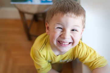 A young boy with short hair sits on the floor and smiles widely. He wears a yellow shirt and looks directly at the viewer with joy. The room is well lit.