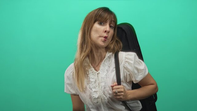 Young woman holding guitar case strap and making puckered lips in a green studio setting; playful travel.