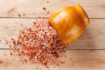 Himalayan salt crystals in grains scattered from a wooden bowl onto a light colored wooden surface.
